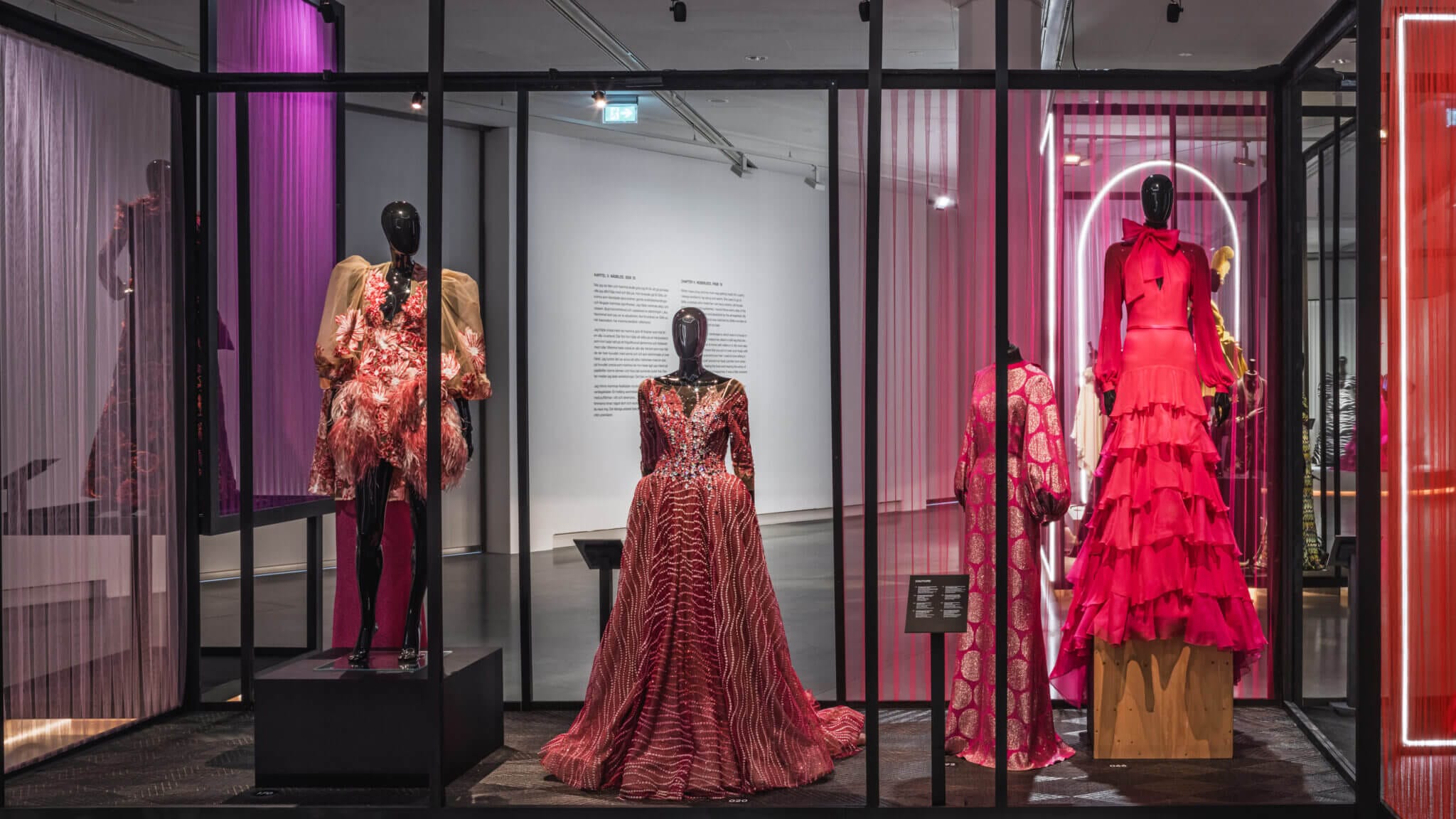 Utställningen visar fem eleganta rosa och röda modeklänningar av Lars Wallin Atelier på skyltdockor i en glasmonter, mot en galleribakgrund med mjuk belysning och förklarande text på väggen.
