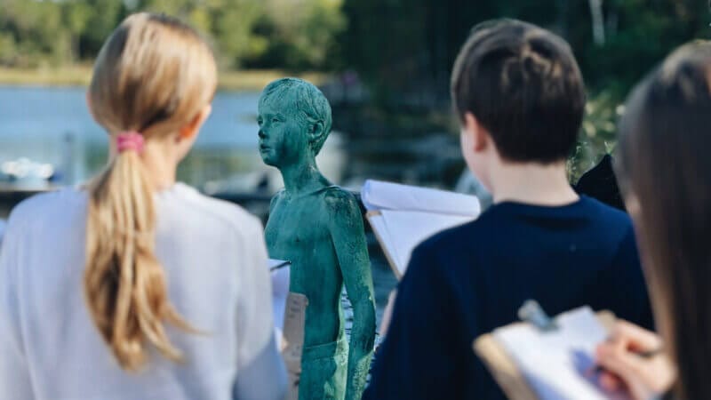 Barn skissar en grön staty av ett barn utomhus nära vatten och håller i skissblock och pennor.