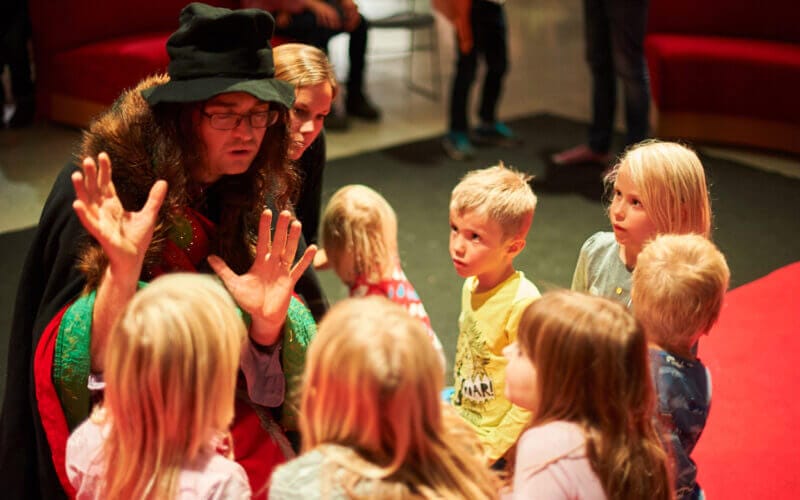 En person i färgglad kostym och hatt pratar livligt med en grupp uppmärksamma barn inomhus.