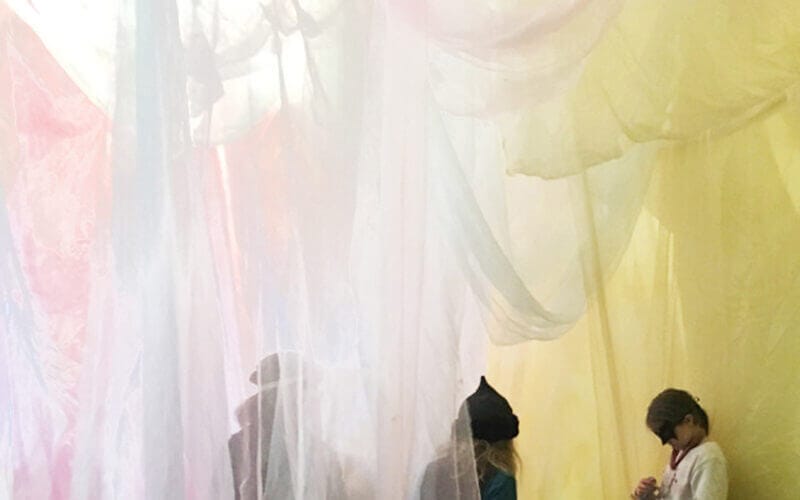 Barn som står bakom skirt, färgglatt tyg som hänger i taket och skapar en mjuk, diffus ljuseffekt.