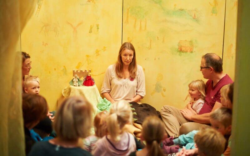 En kvinna läser för en grupp barn och vuxna i ett rum med ljusgula väggar och mjuk belysning. Små dockor och en slottsmodell sitter på bordet bredvid henne.