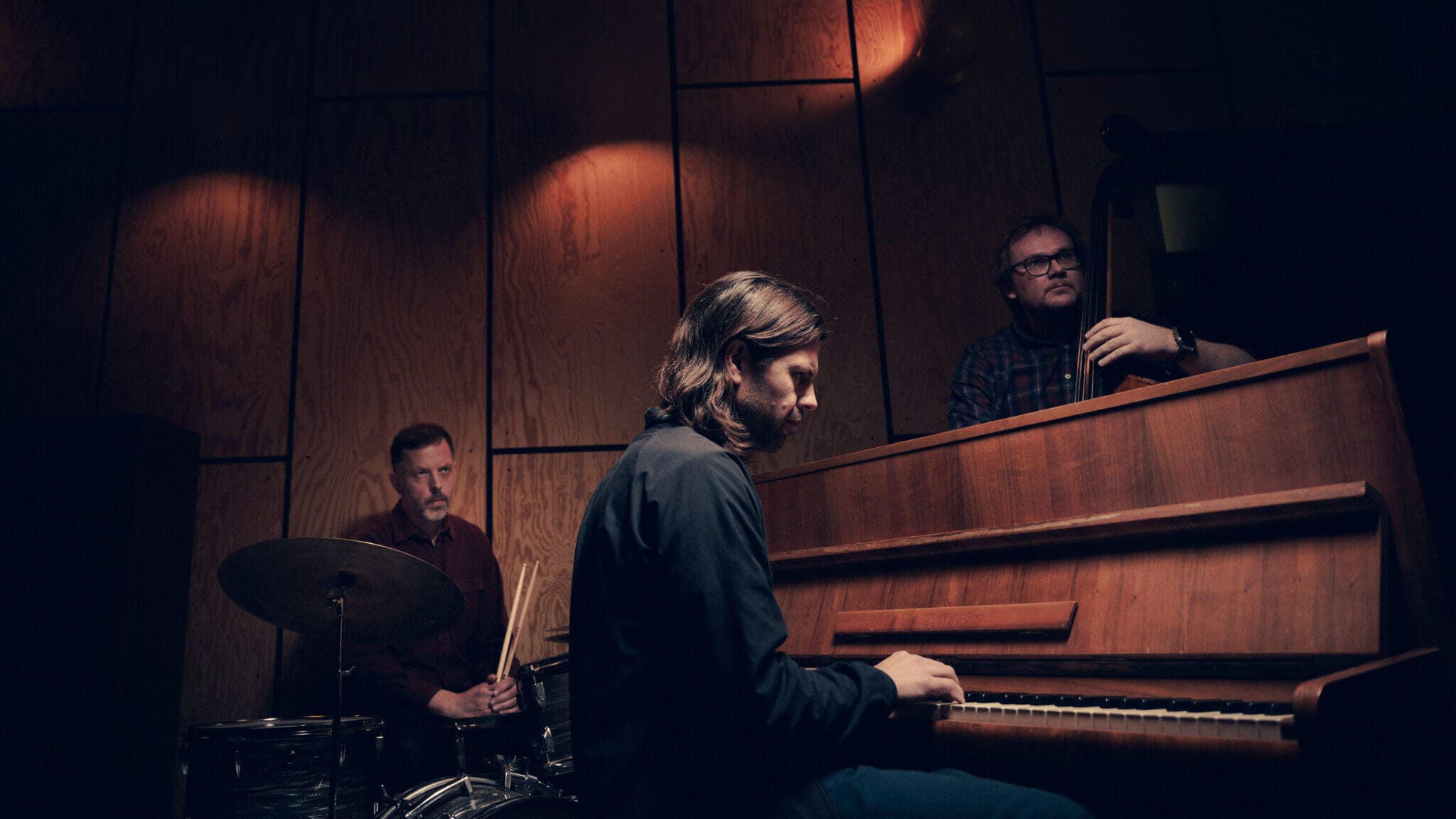 Tre musiker uppträder i ett svagt upplyst rum; denna jazztrio tolkar Bach som en spelar piano, en annan trummor och den tredje står bakom en upprätt bas.