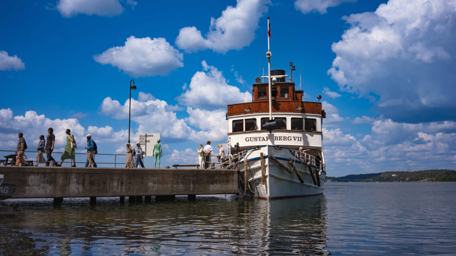 Människor går längs en brygga för att gå ombord på den vita och bruna båten "Gustafsberg VII" en solig dag med spridda moln, på väg mot Artipelag för en naturskön resa.