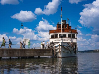 Människor går längs en brygga för att gå ombord på den vita och bruna båten "Gustafsberg VII" en solig dag med spridda moln, på väg mot Artipelag för en naturskön resa.