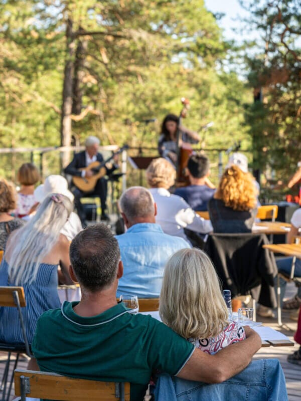 Publiken sitter utomhus på Scensommar 2025 och njuter av ett liveframträdande med gitarr och kontrabas, omgivna av träd i naturligt dagsljus.