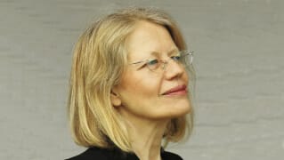 En medelålders kvinna med blont hår och glasögon tittar åt sidan och ler lite, som om hon reflekterar över Den förtrollade skogen - ett divertissement, mot en ljus bakgrund.