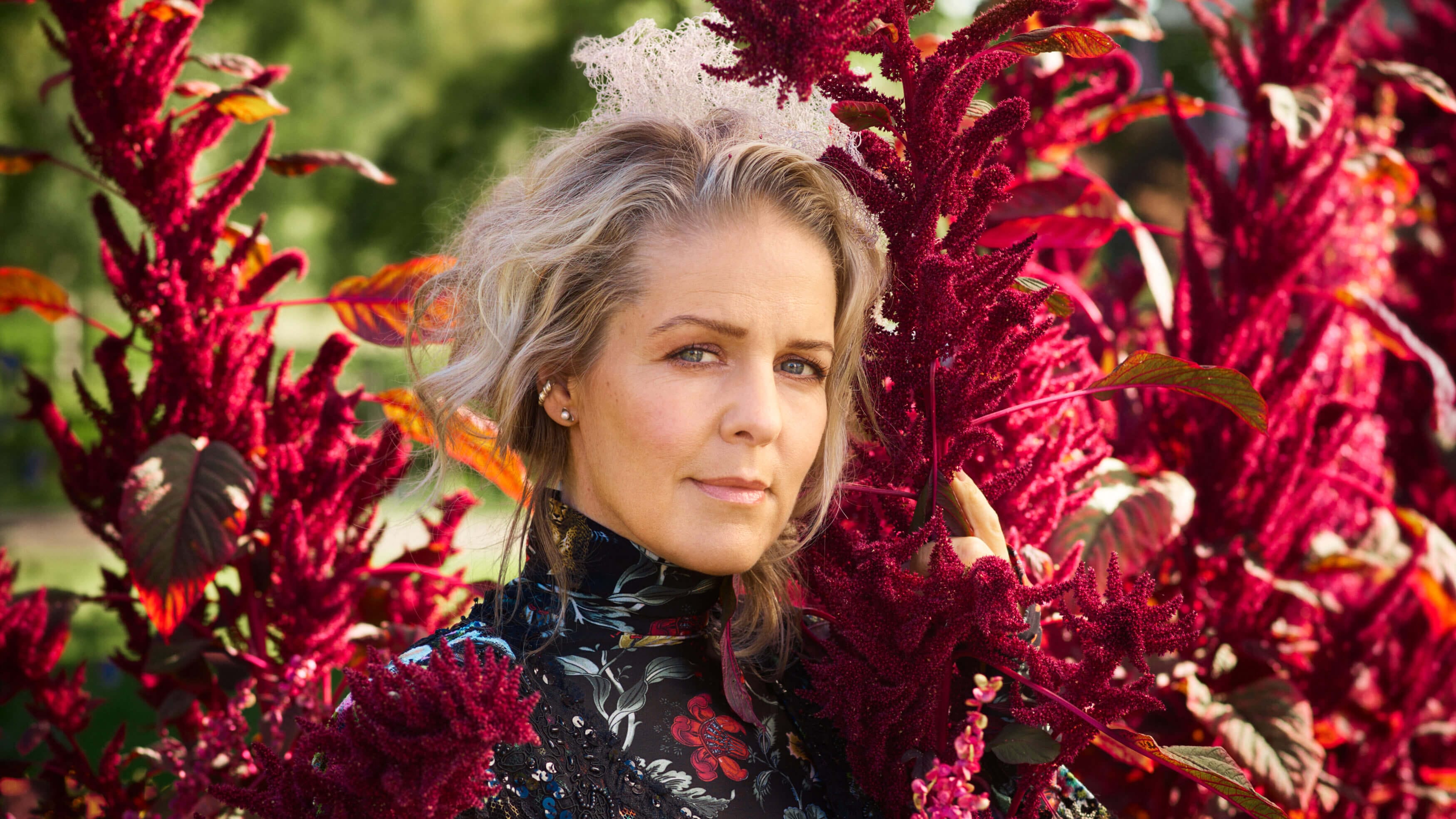 En kvinna med vågigt blont hår och en blommig klänning står bland höga, livfulla magentafärgade blommor utomhus och frammanar den fridfulla andan hos Sofia Karlsson på Artipelag - en sång till Selma, medan hon blickar in i kameran.
