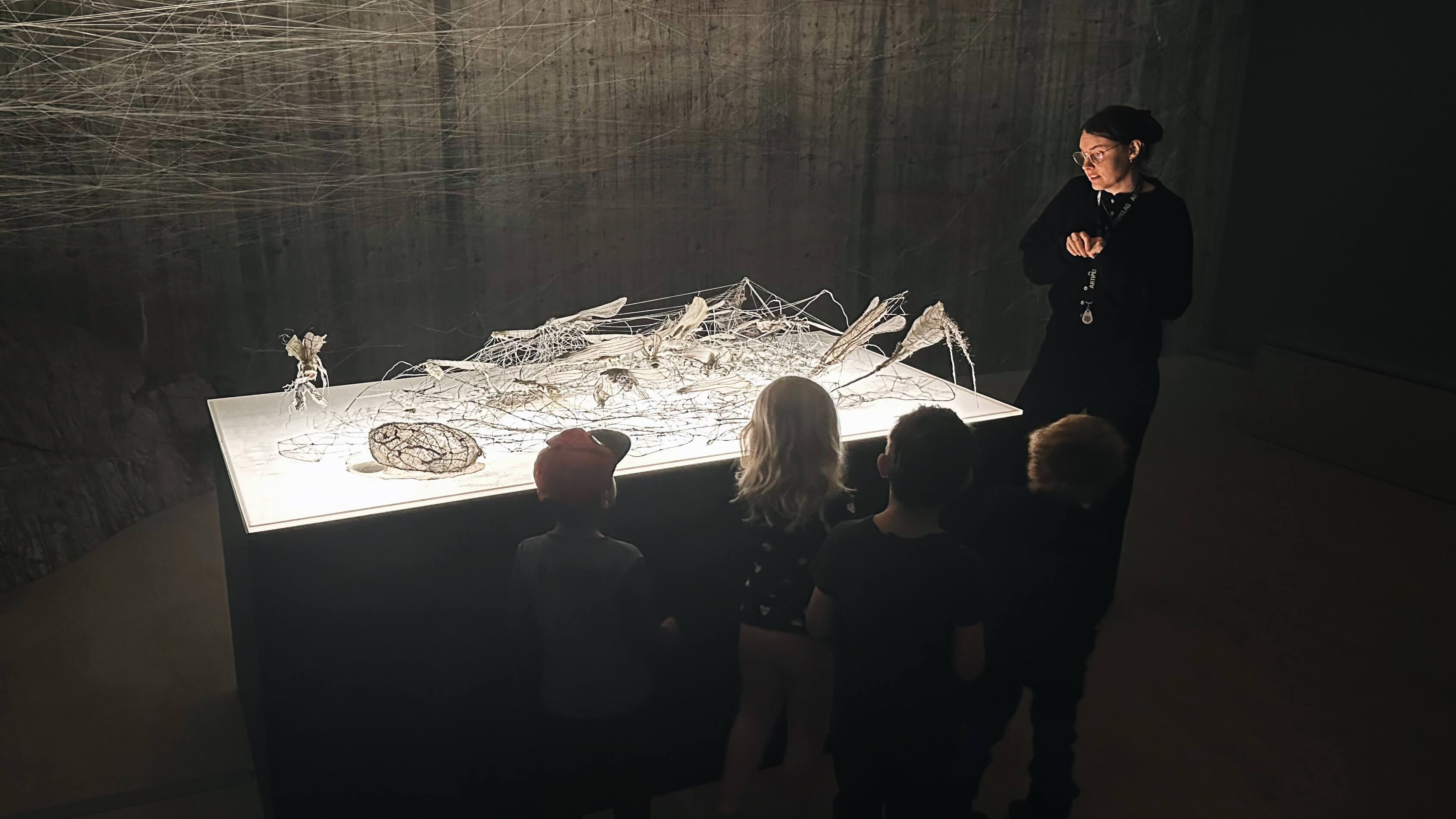 En grupp barn står framför en väl upplyst konstinstallation med metallskulpturer, medan en vuxen förklarar eller observerar i närheten i ett svagt upplyst rum.