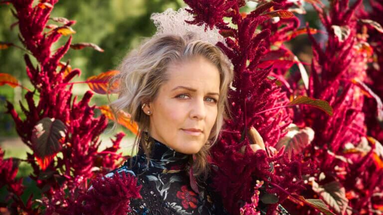 En kvinna med vågigt blont hår och en blommig klänning står bland höga, livfulla magentafärgade blommor utomhus och frammanar den fridfulla andan hos Sofia Karlsson på Artipelag - en sång till Selma, medan hon blickar in i kameran.