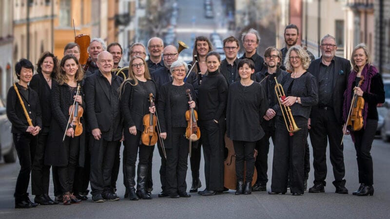 En grupp musiker från Svensk Barock med Drottningholms barockensemble, klädda i svart, står tillsammans utomhus på en gata och håller i olika instrument inklusive fioler, bleckblåsinstrument och en kontrabas.