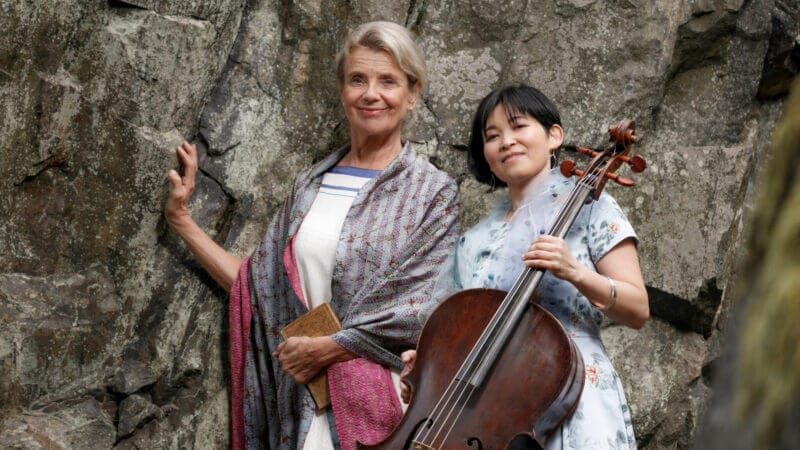 Två kvinnor står framför en klippvägg; den ena håller en bok och bär en sjal, den andra bär en blommig klänning och håller en cello – poesi & musik med Stina Ekblad & Mime Brinkmann.