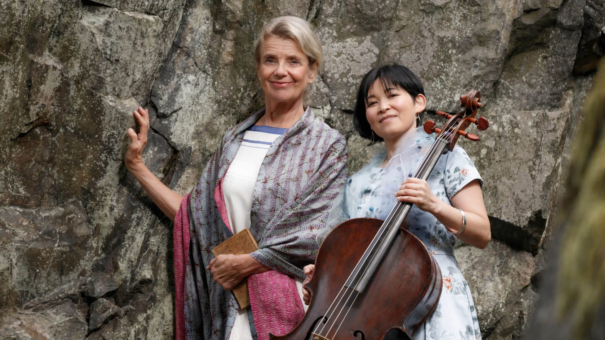 Två kvinnor står framför en klippvägg; den ena håller en bok och bär en sjal, den andra bär en blommig klänning och håller en cello – poesi & musik med Stina Ekblad & Mime Brinkmann.