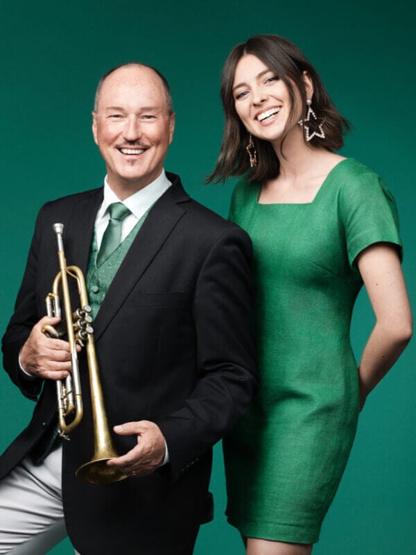 En man i kostym med en trumpet i handen står bredvid en leende kvinna i grön klänning mot en grön bakgrund.
