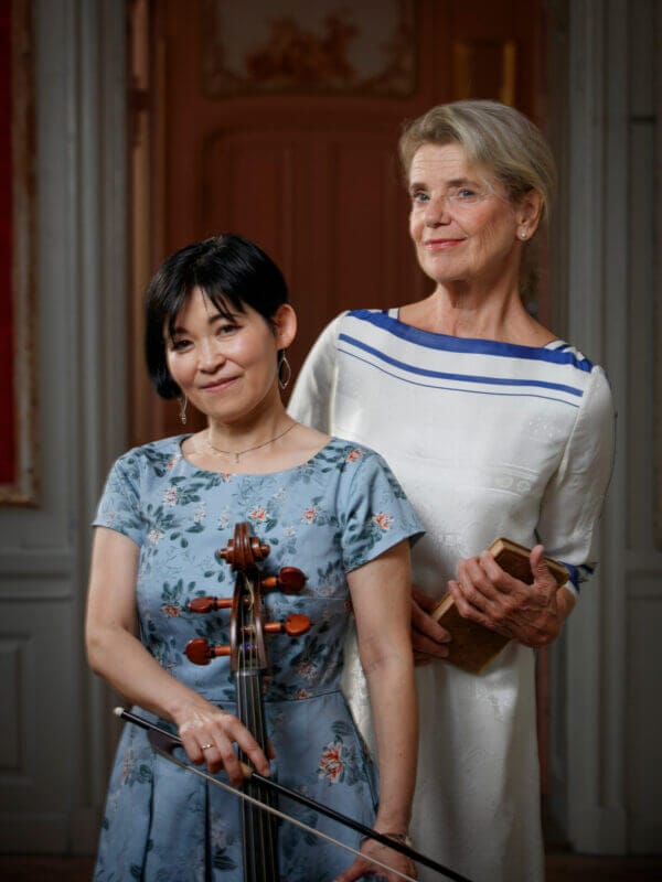 Två kvinnor poserar inomhus; den ena håller en cello, den andra en bok. Stående framför en dörröppning i ljusa klänningar frammanar de andan i "Poesi & musik med Stina Ekblad & Mime Brinkmann".