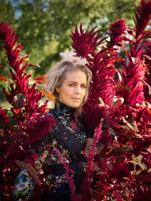 En kvinna med vågigt blont hår och en blommig klänning står bland höga, livfulla magentafärgade blommor utomhus och frammanar den fridfulla andan hos Sofia Karlsson på Artipelag - en sång till Selma, medan hon blickar in i kameran.