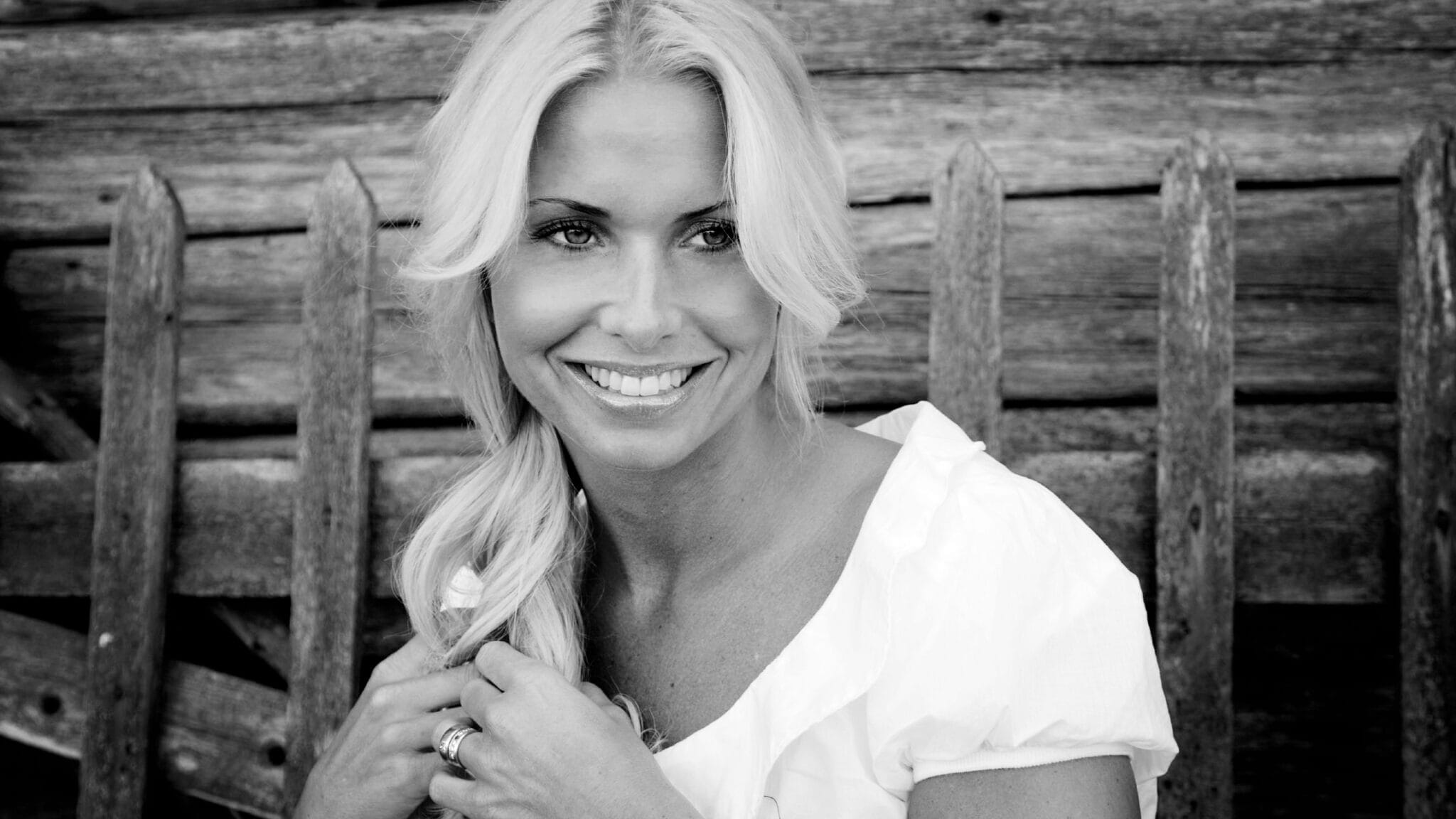 En kvinna med långt blont hår i en vit topp ler medan hon sitter framför ett rustikt trästaket.