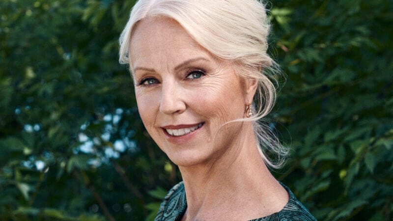 En äldre kvinna med vitt hår uppsatt i en frisyr står utomhus framför gröna bladverk, iklädd en grönmönstrad klänning och ler mot kameran, vilket påminner om Anne Sofie von Otter med vänner – från Piaf till Leander.
