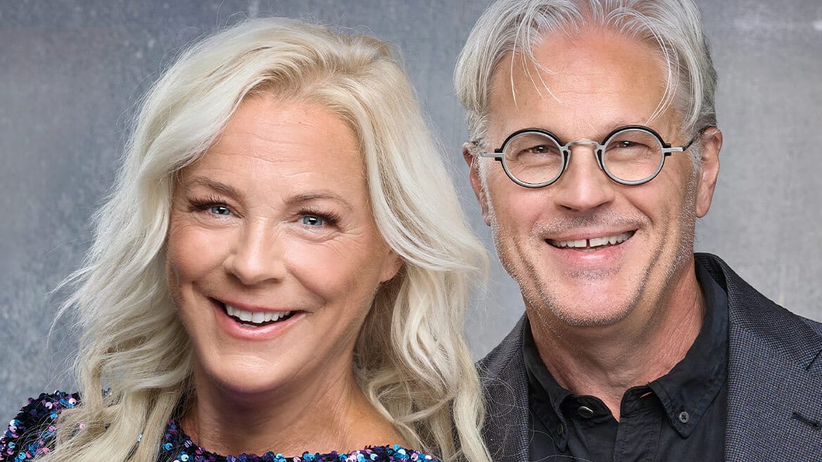 En leende kvinna med långt blont hår står bredvid en leende man med grått hår, glasögon och en mörk jacka mot en neutral bakgrund.