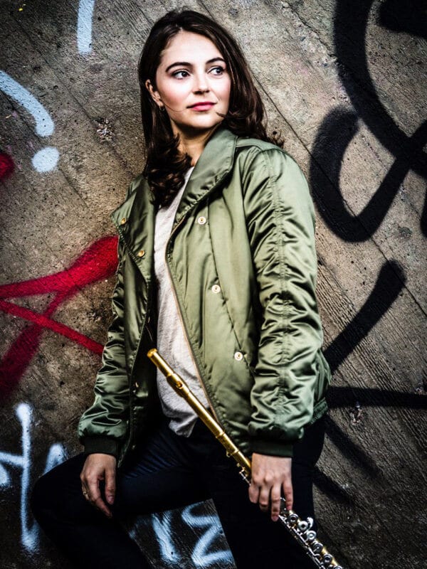En kvinna i grön jacka står mot en graffititäckt vägg och håller en flöjt i silver och guld i sin högra hand – en bild som påminner om svampar – poesi, musik och förvandling.