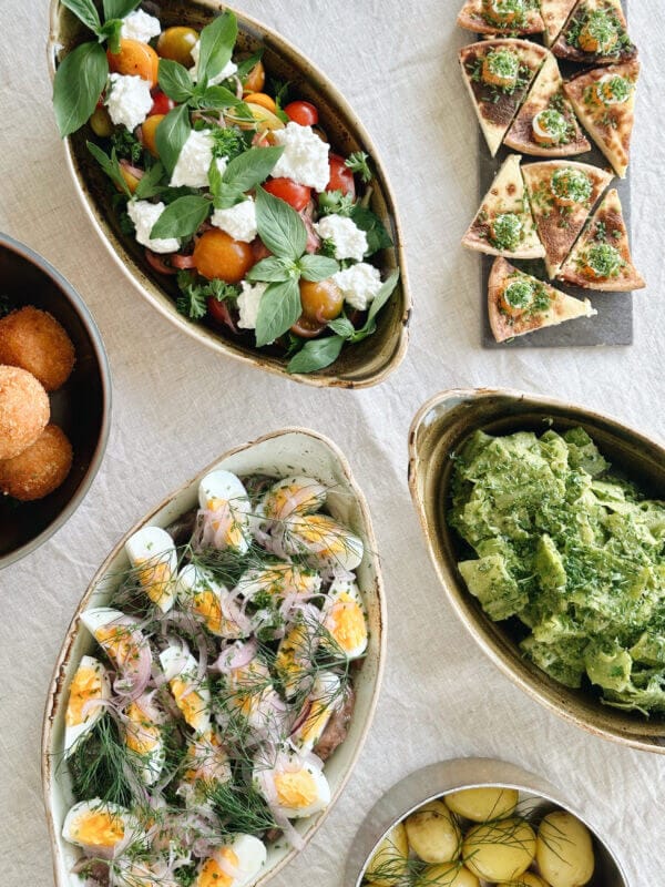 Ett bord med rätter inklusive en tomatsallad med grönsaker, deviled eggs med örter, stekta bollar, grön pasta, örtbaserade flatbrödsskivor och en skål med småpotatis.