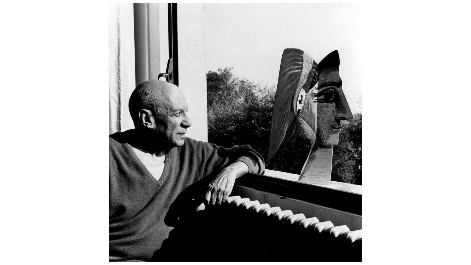 En man sitter vid ett fönster och tittar på en skulpterad byst, med en framträdande näsa, som står på fönsterbrädan; träd syns utanför.