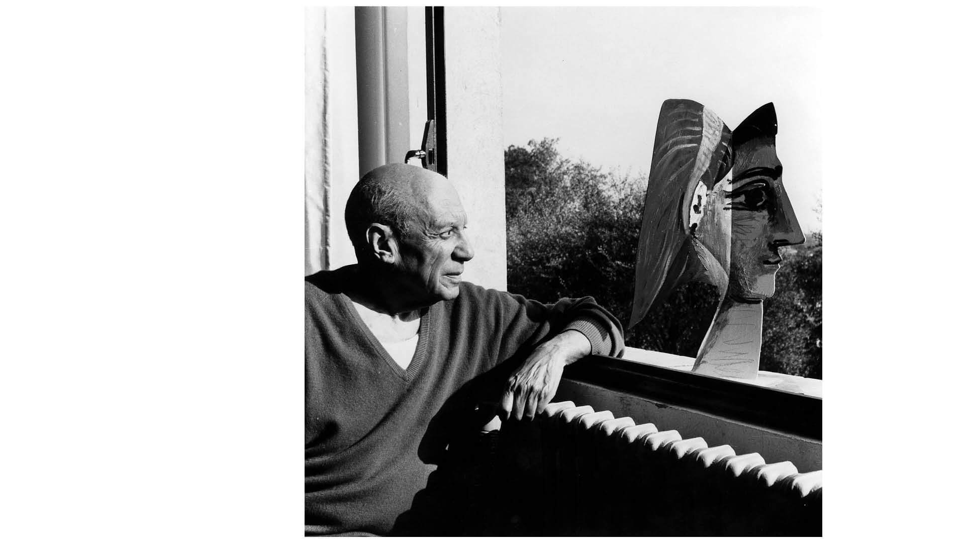 En man sitter vid ett fönster och tittar på en skulpterad byst, med en framträdande näsa, som står på fönsterbrädan; träd syns utanför.