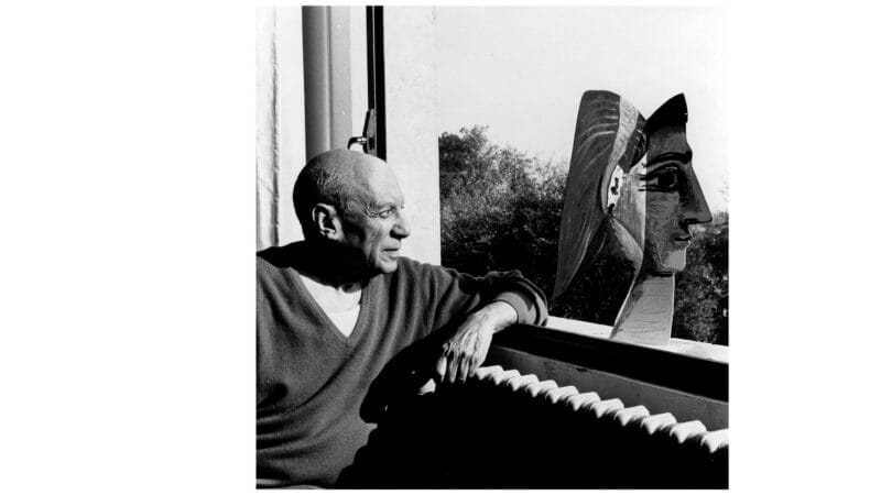 En man sitter vid ett fönster och tittar på en skulpterad byst, med en framträdande näsa, som står på fönsterbrädan; träd syns utanför.