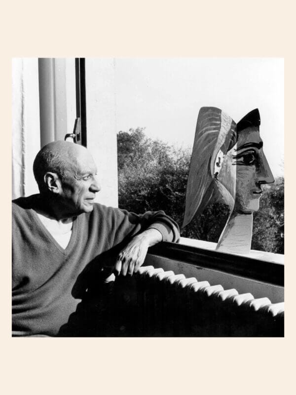 En man sitter vid ett fönster och tittar på en skulpterad byst, med en framträdande näsa, som står på fönsterbrädan; träd syns utanför.