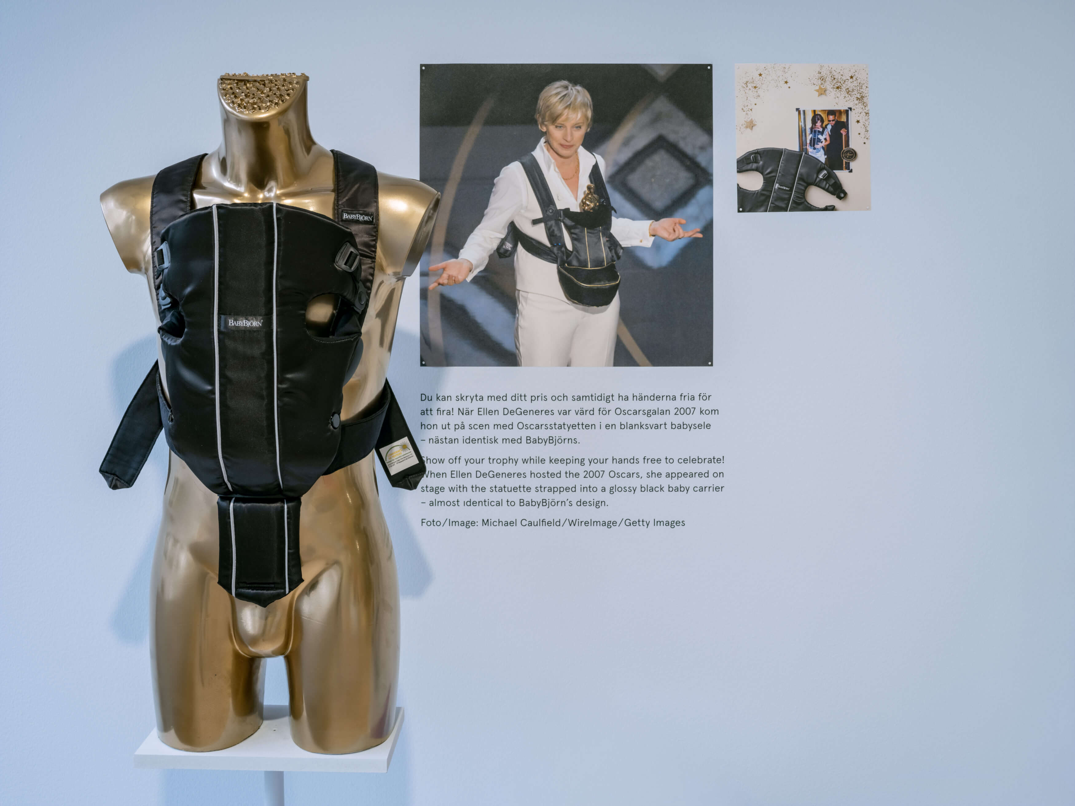 Utställningsbild från utställningen Mitt kreativa liv. Bilden föreställer en docka, som står mot en ljusblå vägg, och som har ett ett svart bärsele. Bredvid på väggen sitter ett fotografi och informationstext.