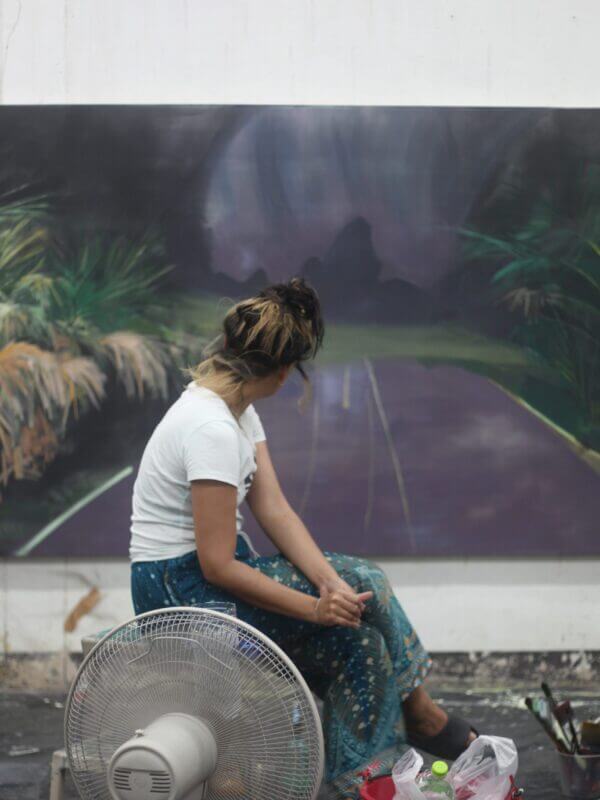 En kvinna som sitter på ett bord och som tittar på en tavla bakom sig. Tavlan föreställer en väg.