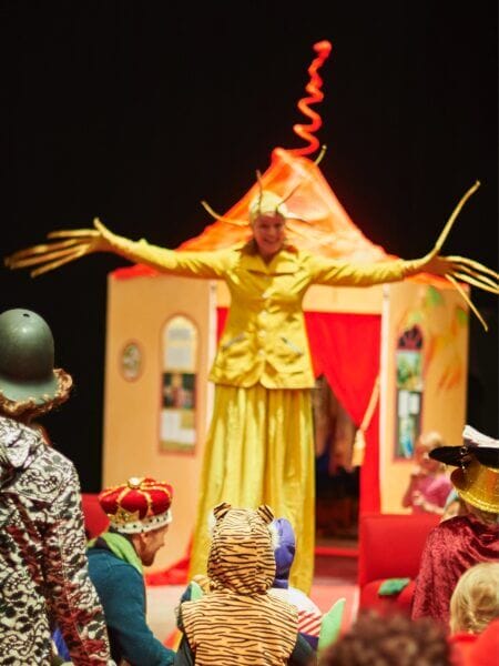 En artist i en gul kostym med långa ärmar står på styltor på en fantasifull scen och underhåller utklädda barn.